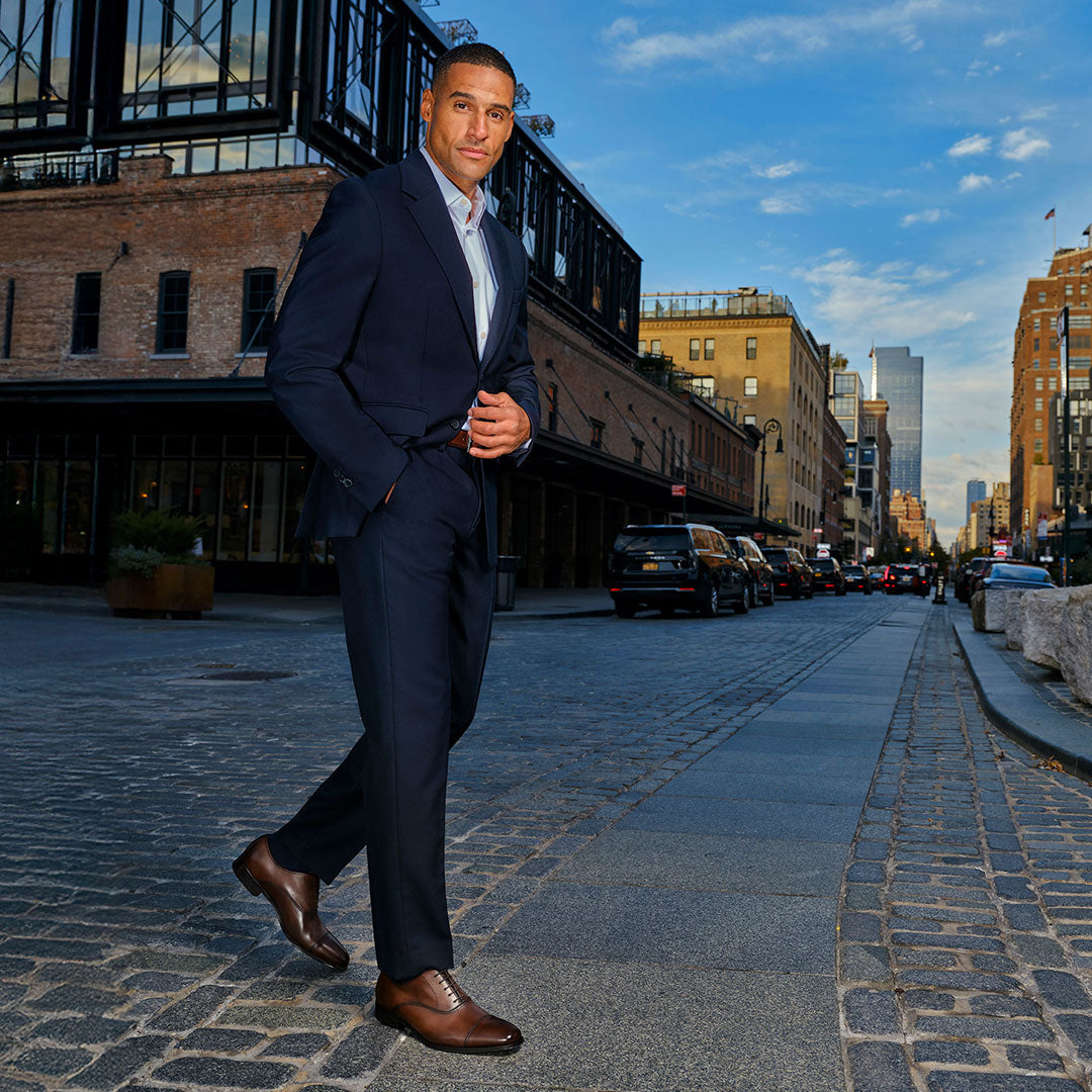 Slide Man in a navy suit wearing To Boot's Aidan Brown Cap Toe Oxford walking on a cobblestone city street with buildings in the background.