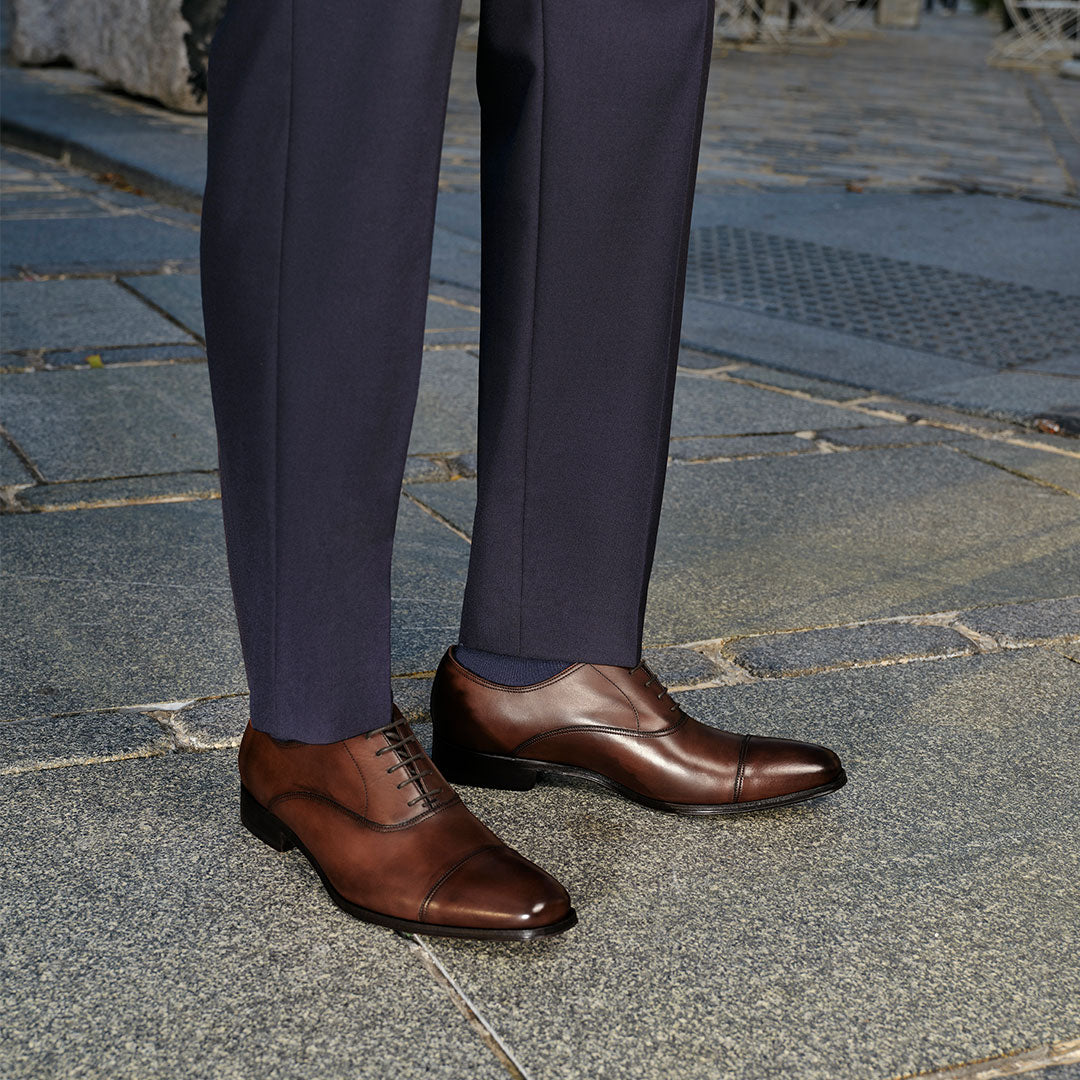 Secondary slide Man in a navy suit pants wearing To Boot's Aidan Brown Cap Toe Oxford standing on a cobblestone city street.