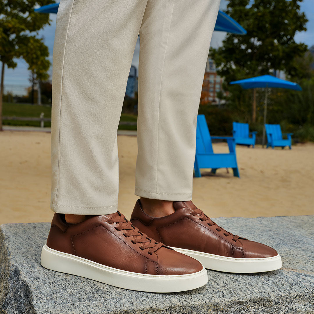 Secondary slide To Boot's James Tan Leather Stretch Lace Luxury Low-Top Sneaker worn with beige pants on a stone surface, with a beach scene in the background.