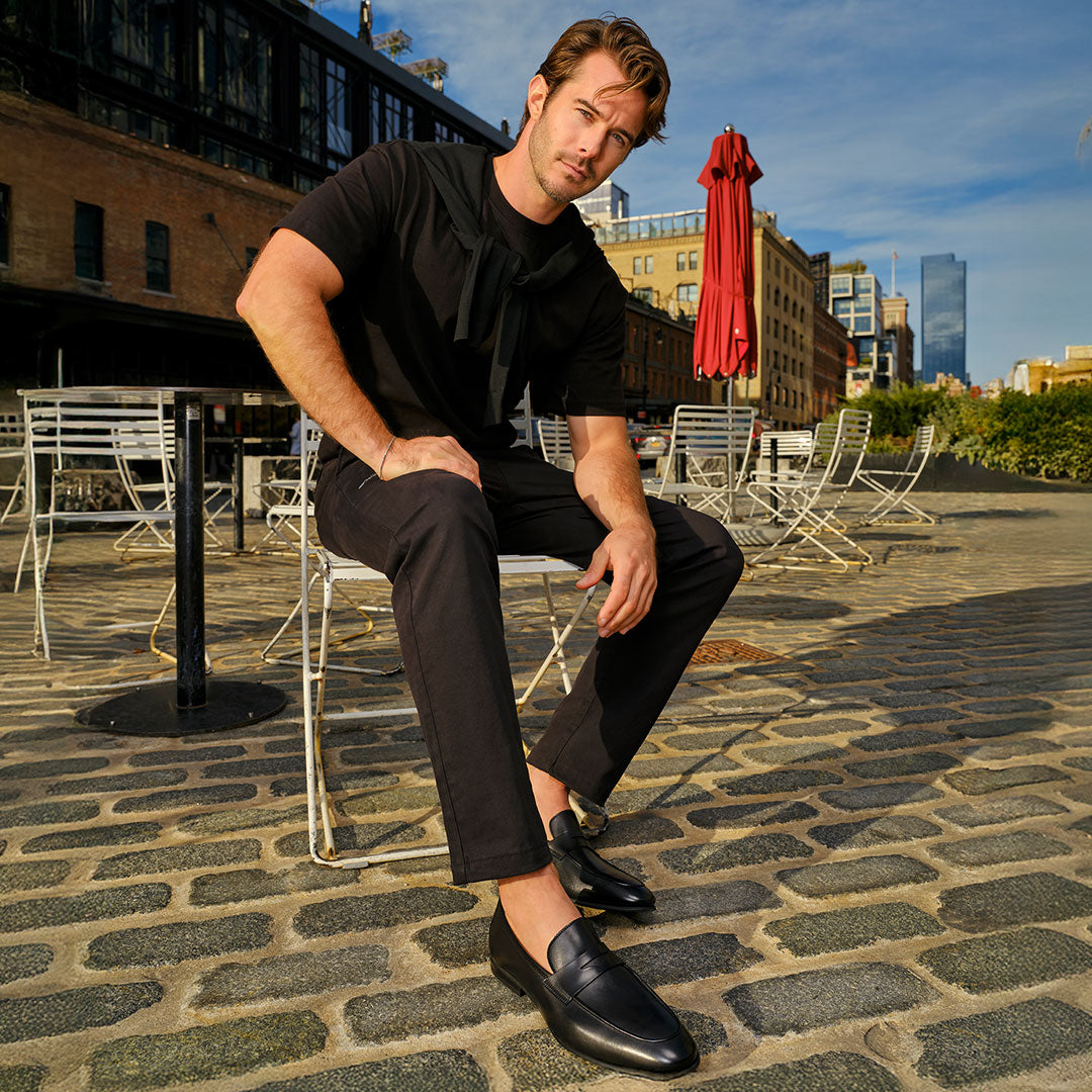 Slide Man in a black outfit wearing To Boot's Keats Black Calf Leather Loafer sitting on a metal chair at an outdoor café on a cobblestone street.